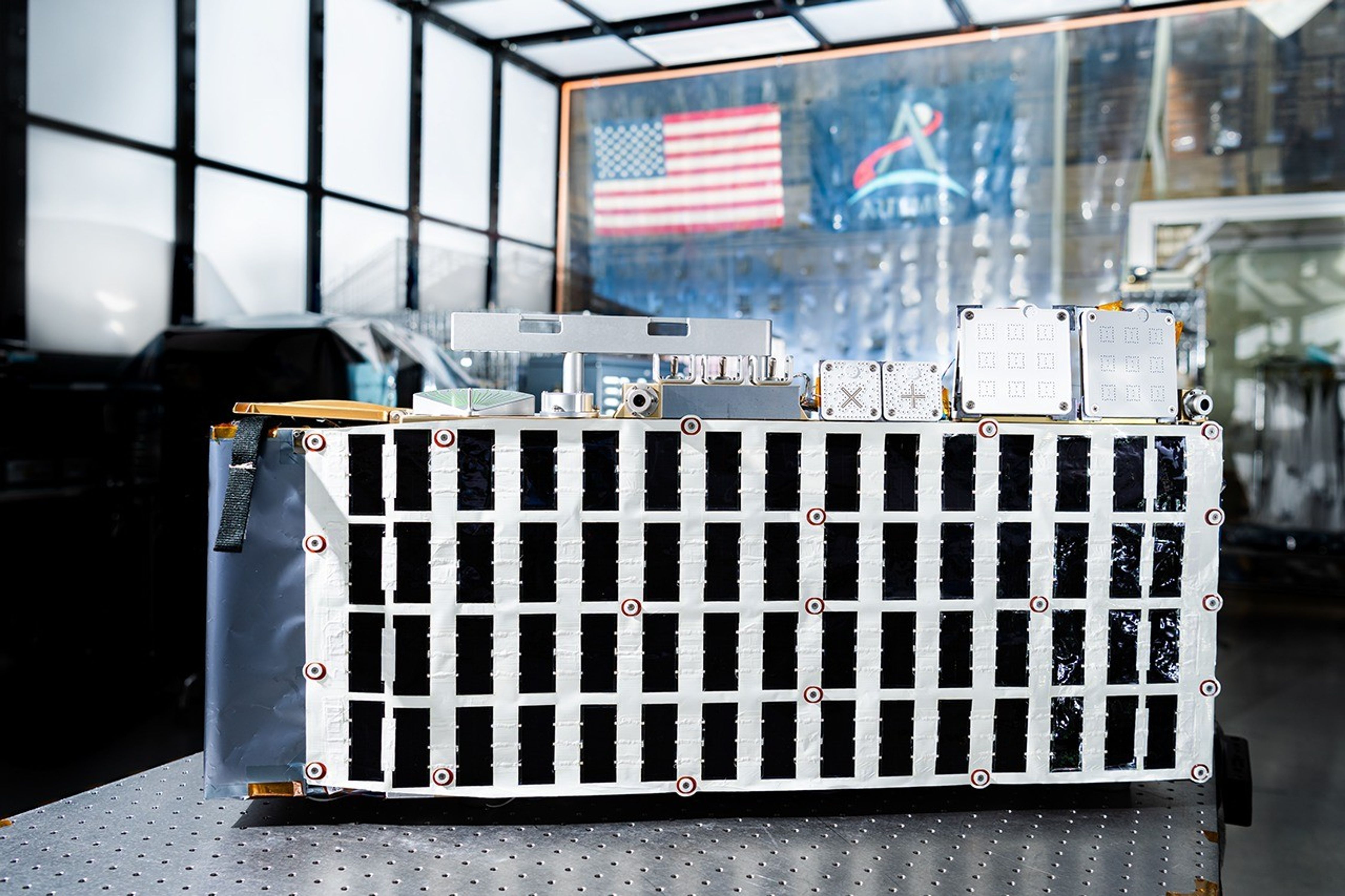 Rectangular box-shaped device resting on a metal table. The front face is covered with a grid of many small black rectangular panels bordered in white. Metal components, brackets, and small box units are mounted along the top. The background shows a large windowed wall with a blurred American flag and an Artemis flag visible behind the device.